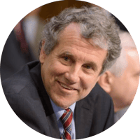 Sherrod Brown, dressed in a suit and tie, smiles leaning forward.